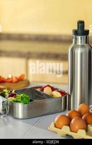 Wiederverwendbares Geschirr aus Metall mit grünem und rotem Blattsalat und rotem Apfel auf einer Holzarbeitsfläche. Eier in einem Karton im Vordergrund, Schneidebrett mit sli Stockfoto