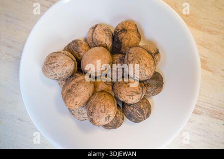 Eine Sammlung ungeschnittener Walnüsse befindet sich in einer weißen Schüssel auf einem Holztisch. Die Nüsse haben eine naturbraune Schale und variieren in Farbe und Textur. Stockfoto