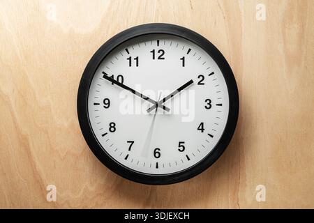 Analoge Wanduhr mit schwarzem Rahmen und weißem Zifferblatt, die an einer hellen Holzwand hängt und die Zeit um 1:50 anzeigt. Ein minimalistisches und klassisches Zeitmesser-Symbol Stockfoto