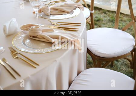 Der wunderschön angeordnete Hochzeitstisch ist mit weichem beigefarbenem Bettzeug, goldfarbenem Besteck und dekorativen Tellern ausgestattet, die das elegante Ambiente für besondere Anlässe unterstreichen Stockfoto