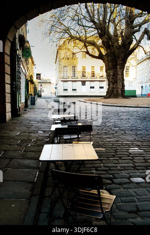 Abbey Green, Bath UK. Leere Tische und Stühle stehen auf einer kopfsteingepflasterten Straße vor Geschäften und einem großen Baum, was auf eine ruhige Nebensaison oder hindeutet Stockfoto