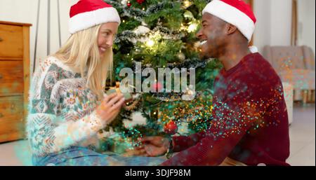 Ein Paar tauscht im Wohnzimmer in der Nähe des dekorierten Weihnachtsbaums ein Geschenk aus, trägt Weihnachtsmützen Stockfoto