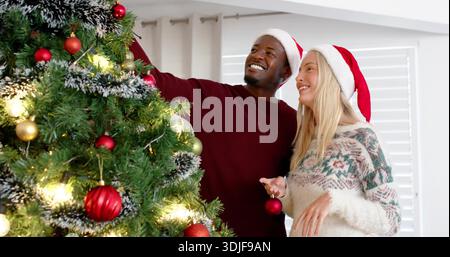 Ein dekoratives Paar, das rote Kugeln auf den Weihnachtsbaum im Wohnzimmer legt, Weihnachtsmützen und Pullover trägt. Urlaub, Ornamente, Lichter, beflockt, sonnenbeleuchtet, gemütlich, Stockfoto