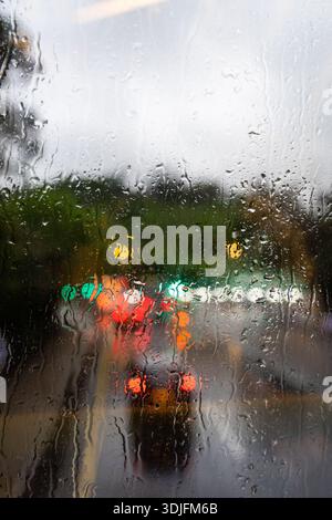 Regentropfen auf Glasfenster. Verschwommene Ampeln im Hintergrund. Stimmungsvolle Atmosphäre. Textur bei nassem Wetter. Filmisches Bokeh. Stockfoto