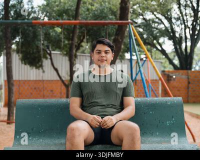 Porträt eines jungen Mannes, der auf einer Bank in einem Stadtpark sitzt und in die Kamera blickt, mit Bäumen und einem unscharfen Kinderspielplatz im Hintergrund. Stockfoto