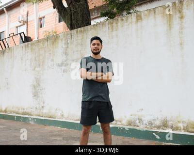 Bild eines jungen Mannes, der mit überkreuzten Armen im Freien vor einer hellen Wand steht. Ideal für die Kommunikation von Selbstvertrauen, urbanen Lebensstil, lässige fas Stockfoto
