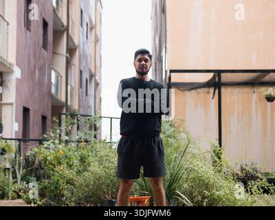 Bild eines jungen Mannes, der mit überkreuzten Armen in einem urbanen Außenraum steht, mit Vegetation und Gebäuden. Perfekt für aktiven Lifestyle Stockfoto