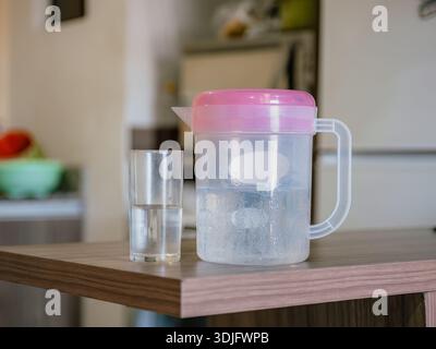 Foto einer Kanne mit Wasser und klarem Glas auf einem Tisch, mit einer unscharfen Küche im Hintergrund. Ideales Bild zur Darstellung der Hydratation, drin Stockfoto