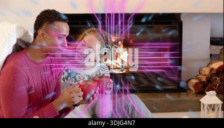 Sitzendes Paar mit rosa cremefarbenen Stricken, die rote Tassen auf der Couch am Kamin halten, lila-blaues Licht Stockfoto