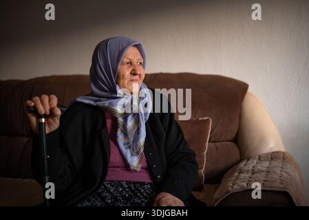 Porträt einer alten muslimischen Frau mit ihrem Gehstock zu Hause, der traditionellen Hijab trägt und wegblickt. Stockfoto