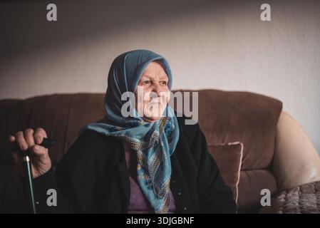 Porträt einer alten muslimischen Frau mit ihrem Gehstock zu Hause, der traditionellen Hijab trägt und wegblickt. Stockfoto