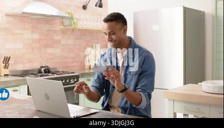 Lächelnder Mann mit Denim-Hemd und Armbanduhr, der in der modernen Küche einen Laptop mit Kreditkarte abklopfen kann Stockfoto