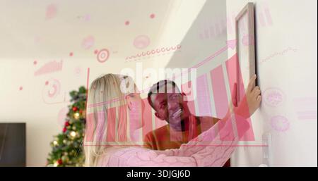 Hängendes Paar, das gerahmtes Bild an der Wand am Weihnachtsbaum ausrichtet, Frau in rosa Strick, rosa Karten Stockfoto