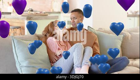 Kuschelndes Paar mit rostbraunen und rosafarbenen Pullover auf dem Sofa im Wohnzimmer, mit Herzballons Stockfoto