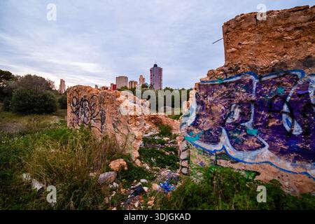 Ein Profilblick auf verlassene Steinmauern mit geschichteten Graffiti, mit den modernen Türmen von Benidorm, die im sanften Abendlicht dahinter aufsteigen. Stockfoto