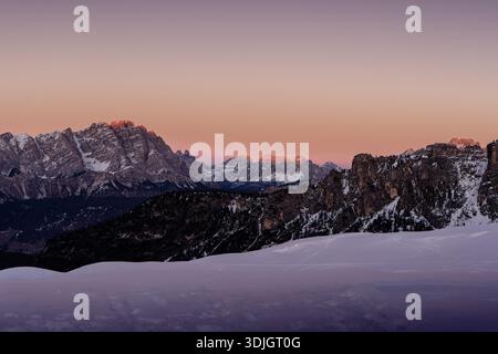 Wintersonnenuntergang am Passo Giau in den Dolomiten, wo warme alpine Lichter mit schneebedeckten Gipfeln unter einem dramatischen violetten Himmel kontrastieren. Stockfoto