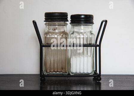 Zwei Salz- und Pfefferstreuer aus Glas in einem Metallhalter auf einem dunklen Tisch vor neutralem Hintergrund. Minimales Detail in der Küche. Stockfoto