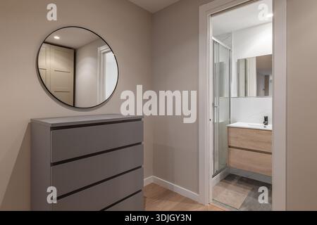 Modernes Badezimmer mit grauer Kommode und rundem Spiegel. Der Raum ist gut beleuchtet und verfügt über eine Glasdusche und einen Holzwaschtisch. Stockfoto