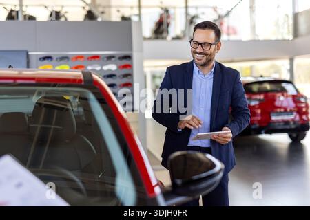 Abgeschnittenes Porträt eines hübschen jungen männlichen Autoverkäufers, der auf dem Ausstellungsboden arbeitet. Glücklicher Verkäufer, der in einem Autohaus steht und in die Kamera blickt. Stockfoto