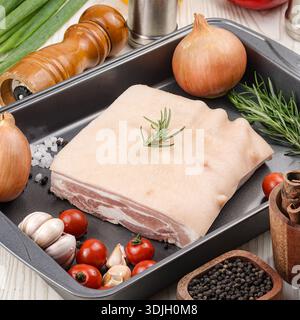Ein Stück roher Schweinebauch mit Rinde wird in ein dunkles Backblech gelegt, umgeben von frischen Zutaten wie Rosmarinzweigen, Zwiebeln, Knoblauch und Kirschtoma Stockfoto