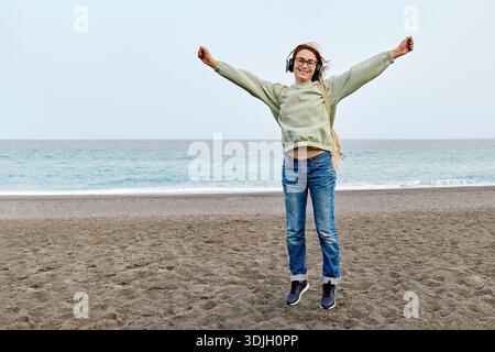 Lächelnde Frau mittleren Alters im Kopfhörer, die Musik hört und tanzt, während sie in der Nähe des Meeres spaziert. Stockfoto