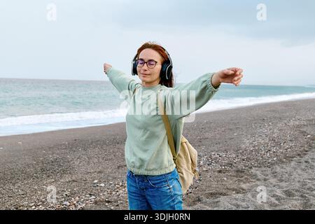Lächelnde Frau mittleren Alters im Kopfhörer, die Musik hört und tanzt, während sie in der Nähe des Meeres spaziert. Stockfoto