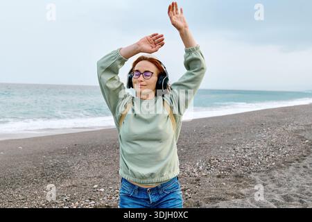 Lächelnde Frau mittleren Alters im Kopfhörer, die Musik hört und tanzt, während sie in der Nähe des Meeres spaziert. Stockfoto
