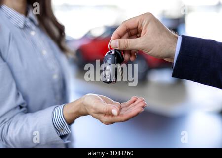 Eine junge weiße Frau kauft ein Auto in einem Autohaus. Der Verkäufer gibt die Autoschlüssel. Lächelnder Verkäufer oder Manager, der der modernen glücklichen Frau, die neu ist, Autoschlüssel gibt Stockfoto