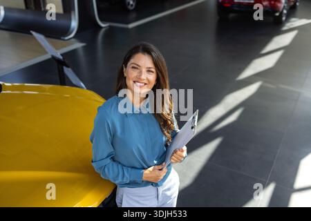 Lächelnde Verkäuferin, die einen Tablet-PC hält, während sie die Kamera im Ausstellungsraum eines neuen Autos betrachtet. Professioneller Autohändler posiert stolz im Autohaus und lächelt zu Stockfoto