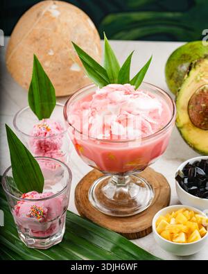 Rosafarbenes Dessert mit Früchten und Blättern, serviert in Glasschalen auf weiß mit verschiedenen Belägen und tropischen Blättern. Stockfoto