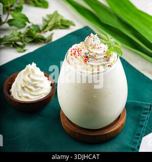 Ein dekadenter Vanillemilkshake, serviert in einem runden Glas, gekrönt mit Schlagsahne, bunten Streuseln und einem frischen Minzblatt. Ein kleines Holz Stockfoto