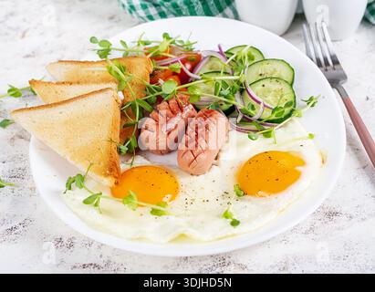 Köstliches Frühstück mit Spiegeleiern, gegrillten Würstchen, Tomaten, Gurke, Gemüse und Gewürzen auf einem Teller auf hellem Hintergrund. Stockfoto