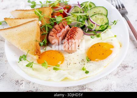 Köstliches Frühstück mit Spiegeleiern, gegrillten Würstchen, Tomaten, Gurke, Gemüse und Gewürzen auf einem Teller auf hellem Hintergrund. Stockfoto
