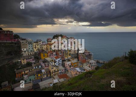 Der Blick auf farbenfrohe Gebäude stürzt unter einem dramatischen, wolkenreichen Himmel zum Meer hinunter und bildet einen lebhaften Kontrast zum tiefblauen Wasser Manaro Stockfoto