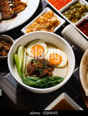 Eine gemütliche Schüssel mit herzhaftem Congee, einem traditionellen Reisbrei, wird mit perfekt weich gekochten Eiern, gewürztem Hackfleisch und frischem grünen Gemüse garniert Stockfoto