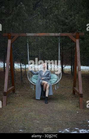 Frau sitzt auf einer Schaukel in einer Waldlichtung. Sie sieht entspannt aus, während sie das Nachmittagslicht genießt. Schnee ist auf dem Boden zwischen den Bäumen zu sehen. Stockfoto