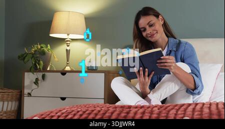 Liest eine Frau im mittleren Erwachsenenalter auf dem Schlafzimmerbett, trägt ein Denim-Hemd mit marineblauer Hardcover, schwebende Bits Stockfoto
