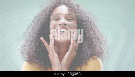Lächelnde Frau berührt das Gesicht im Studio, trägt Senftop mit Ringen, Halskette und digitalen Wirbeln Stockfoto