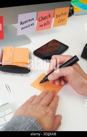 Ein Mann legt eine Hand auf einen Schreibtisch, während er einen schwarzen Marker hält, um auf eine leere orangefarbene Haftnotiz zu schreiben, mit Büroklammern, einem Smartphone und Aufgabe nicht Stockfoto