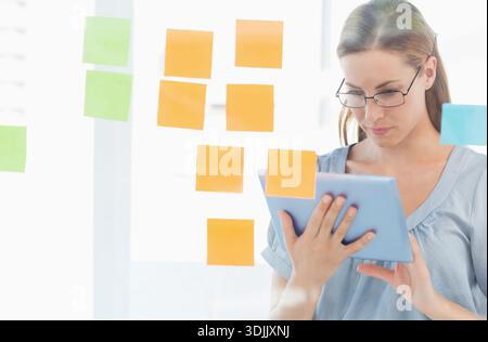 Blaue Bluse für Frauen im mittleren Alter, Gläser mit Tablette, Markierungsnotizen auf Glas im Büro, Kopierraum. Planung, Brainstorming, Fokus, Gerät, Hintergrundbeleuchtung, AA Stockfoto