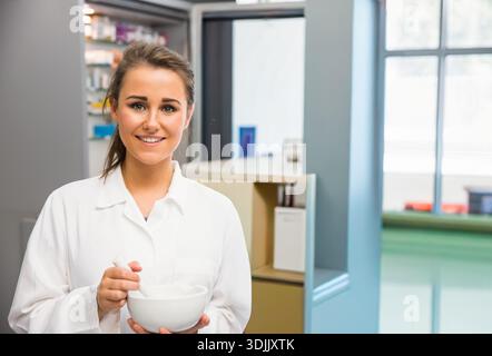 Frau Mitte 20 mit weißem Laborkittel, der Mörtel und Stößel in der Apotheke hält und umrührt. Apotheke, Gesundheitswesen, medizinisch, klinisch, Tageslicht, Frien Stockfoto
