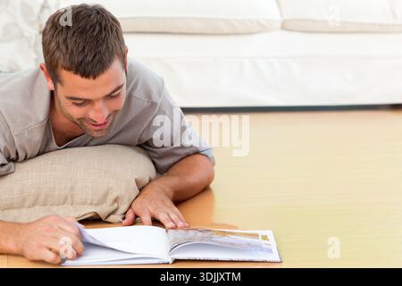 Mann, der liegend auf Holzboden im Wohnzimmer liegt, sich auf beigefarbenem Kissen stützt, Magazinblätter dreht. Leser, Heim, Sofa, Kissen, Lounge, entspannt, sonnig Stockfoto