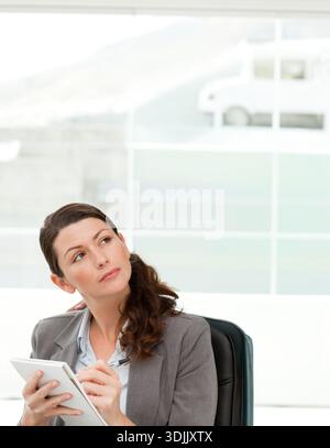 Frau in den 30ern, die im Büro in einem schwarzen Lederstuhl sitzt und einen Blazer trägt, Notizblock, Stift und Blick hält. Weiblich, professionell, Tablet, Stift, Arbeitsplatz, da Stockfoto