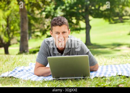 Mann, der im grauen Polo auf dem Bauch liegt, mit silbernem Laptop auf blau-weiß karierter Decke im Park. Im Freien, Natur, Gras, Bäume, Sonnenlicht, lässig, Rela Stockfoto