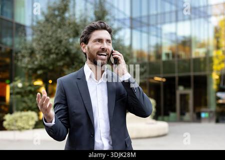 Glücklicher Geschäftsmann, der draußen steht und ein Telefongespräch führt, in eleganter Geschäftskleidung Freude und Kommunikation ausdrückt Stockfoto