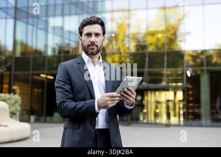 Selbstbewusster bärtiger Geschäftsmann, der draußen vor einem modernen Glasbürogebäude steht, ein digitales Tablet hält und mit ihm interagiert, was die Integration von Technologie in das Berufsleben repräsentiert Stockfoto
