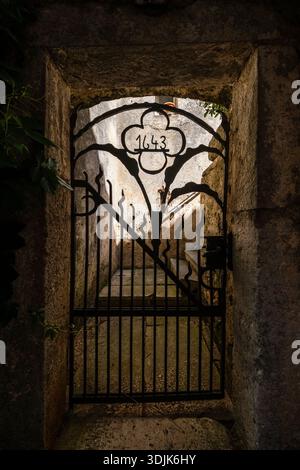 Reich verziertes schmiedeeisernes Tor mit dem Jahr 1643 in einem Steinbogen. Schmale Steinstufen sind in der Entfernung bei weichem Licht sichtbar. Stockfoto