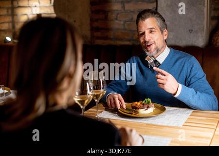 Ein Mann sitzt an einem Holztisch in einem Restaurant. Er lächelt, während er seine Mahlzeit isst. Eine Frau sitzt ihm gegenüber und beobachtet ihn. Beide haben Gläser Wein n Stockfoto