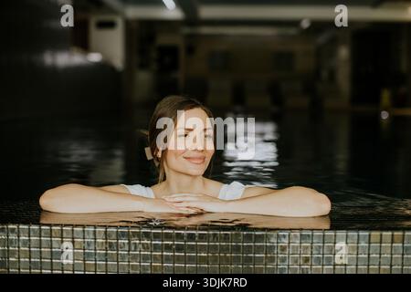 Eine Frau entspannt sich in einem Schwimmbad mit den Armen am Rand. Sie lächelt und genießt die ruhige Atmosphäre des Innenraums, während das Wasser umgibt Stockfoto