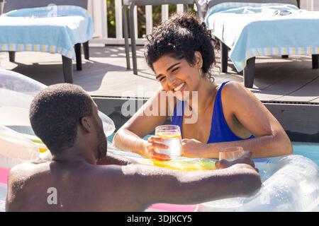 Diverse Paare in Badeanzügen plaudern, lehnen sich auf aufblasbare am sonnigen Hinterhof-Pool und halten Getränke Stockfoto